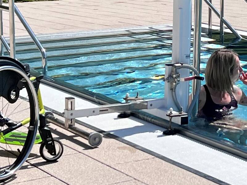 Auf dem Bild wird eine Frau mit dem Lift ins Freibad gebracht. Der Rollstuhl steht am Beckenrand. 