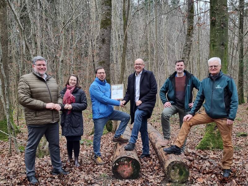 Sechs Personen stehen im herbstlichen Wald auf Laub und posieren für ein Foto, während zwei von ihnen ein Dokument in der Hand halten.