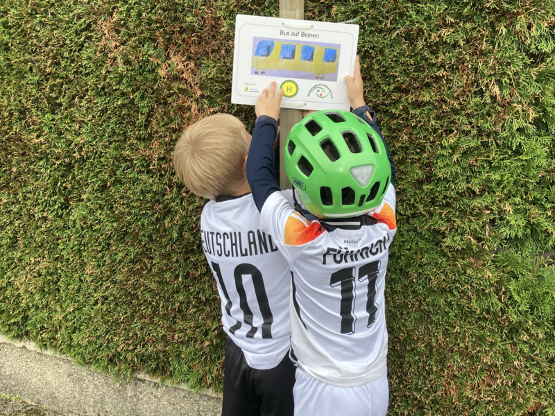 Zwei Kinder in Fußballtrikots betrachten ein Schild an einem Holzpfosten vor einer grünen Hecke.