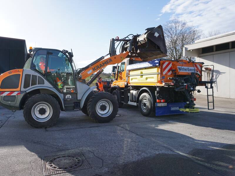 Ein Bagger lädt Material in einen orangefarbenen Winterdienst-Lkw.