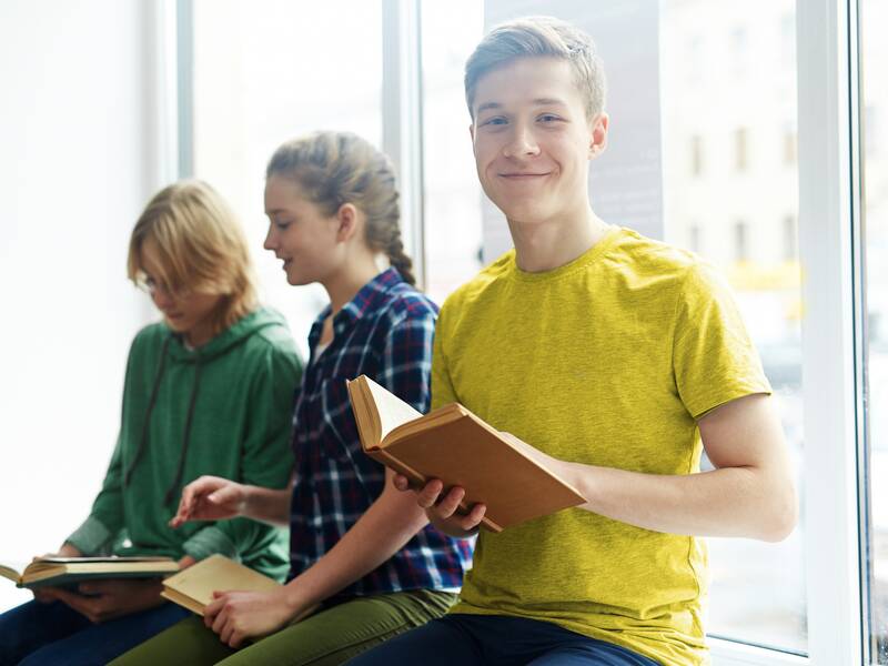 Ein junge sitzt mit einem offenen Buch neben zwei Mädchen an einem Fenster.