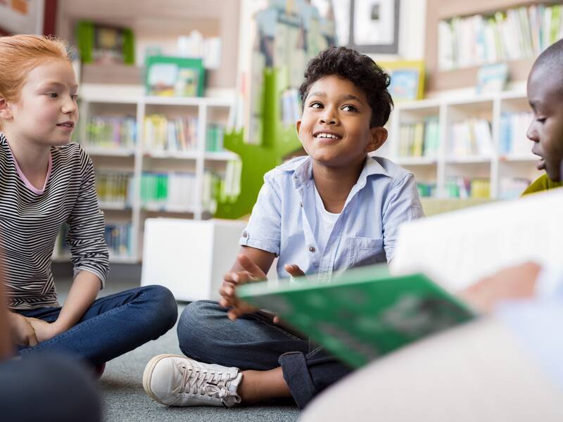 Zwei kleine Jungen und ein Mädchen sitzen auf dem Boden. Im Vordergrund hält eine Person ein Buch in den Händen.