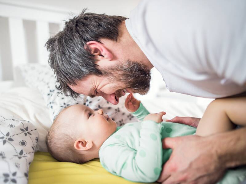 Ein Vater beugt sich zu seinem Baby, dass auf einem Wickeltisch oder einem Bett liegt.