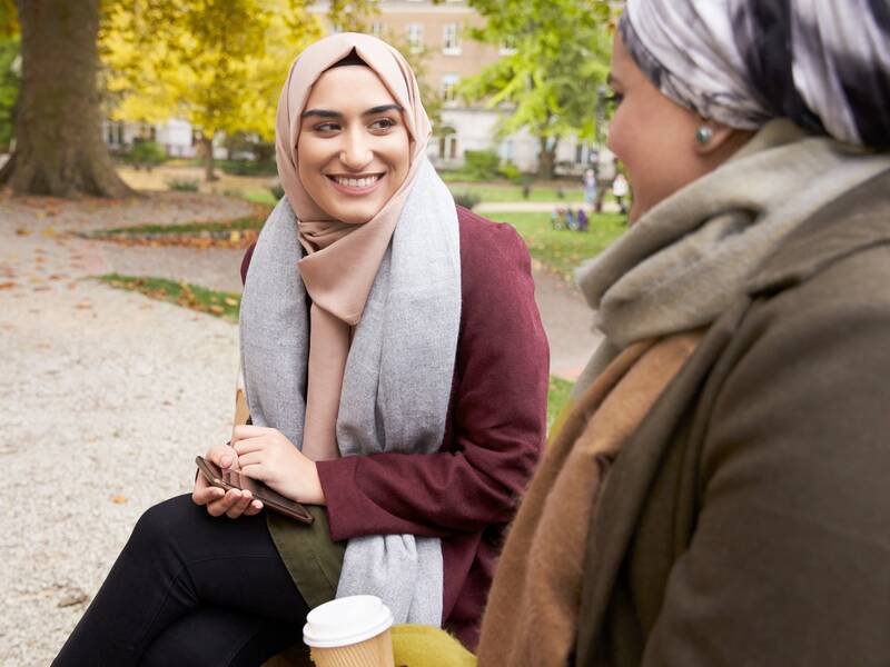 Zwei junge Frauen mit Kopftuch sitzen auf einer Bank im Park und unterhalten sich.