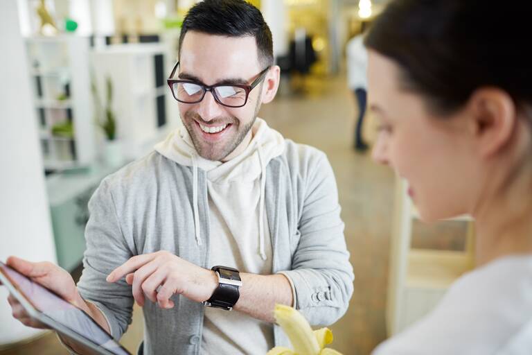 Ein junger Mann mit Brille hält ein Tablet in der rechten Hand und zeigt mit der linken Hand darauf. Rechts daneben steht eine Frau.