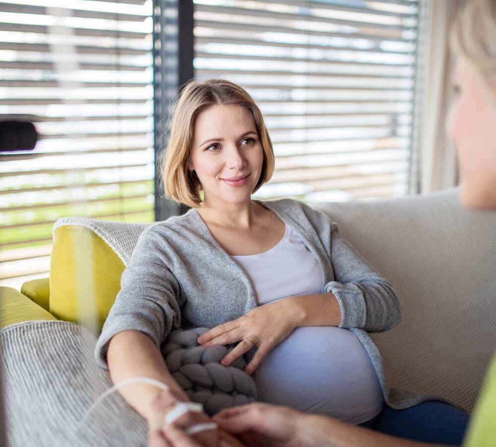 Eine schwangere Frau sitzt auf einem Sofa. In ihrer rechten Hand steckt einen Nadel mit einer Infusion. Rechts im Bild ist eine Frau, die die Hand der Schwangeren hält.