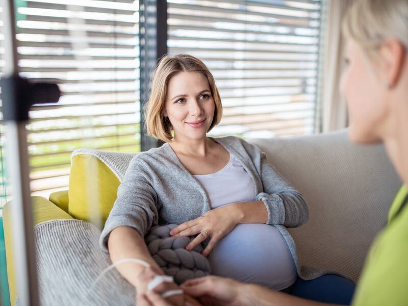 Eine schwangere Frau sitzt auf einem Sofa. In ihrer rechten Hand steckt einen Nadel mit einer Infusion. Rechts im Bild ist eine Frau, die die Hand der Schwangeren hält.