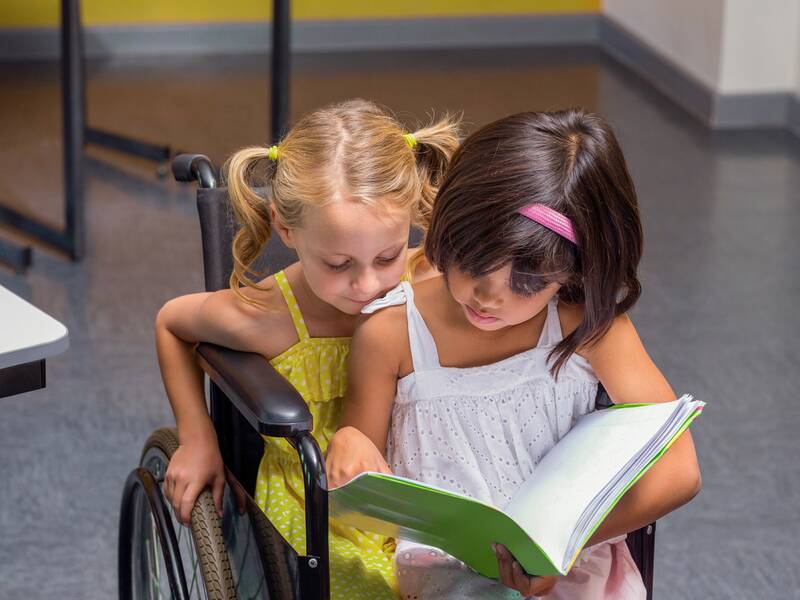 Zwei kleine Mädchen sitzen zusammen in einem Rollstuhl und schauen in ein Buch.