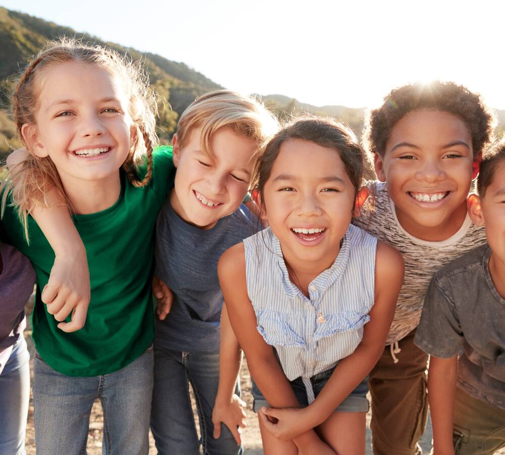 Drei Mädchen und drei Jungs stehen vor einem hügeligen Hintergrund und lachen. Sie halten sich an den Schultern fest.