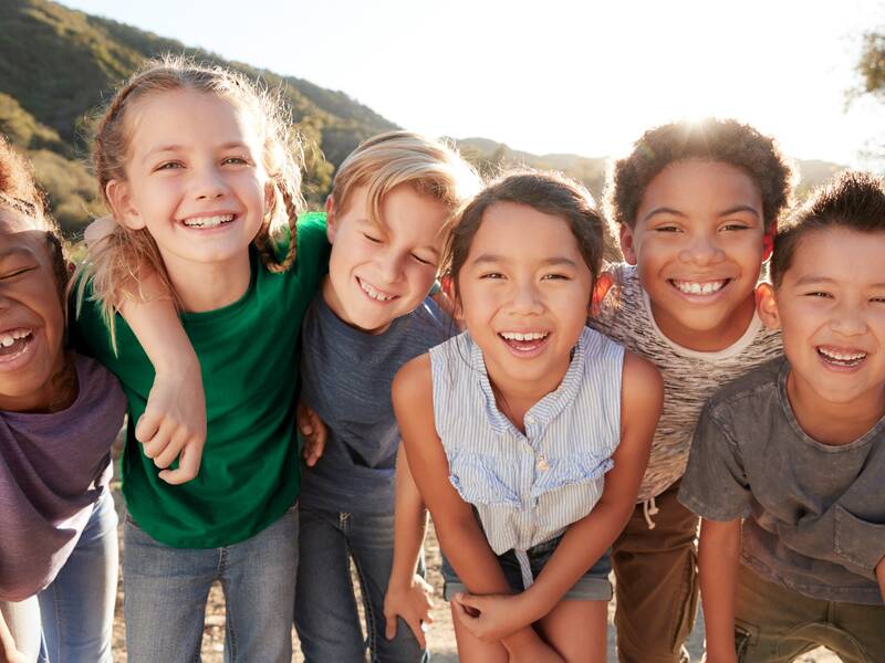 Drei Mädchen und drei Jungs stehen vor einem hügeligen Hintergrund und lachen. Sie halten sich an den Schultern fest.