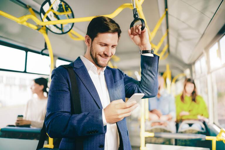 Ein Mann in einem Anzug steht in einem Bus und hält sich mit der linken Hand an einem Haltegriff fest. In der rechten Hand hält er ein Smartphone, auf das er schaut. Im Hintergrund sitzen weitere Passagiere im Bus.