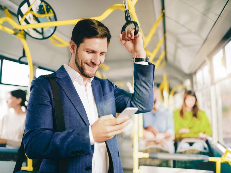 Ein Mann in einem Anzug steht in einem Bus und hält sich mit der linken Hand an einem Haltegriff fest. In der rechten Hand hält er ein Smartphone, auf das er schaut. Im Hintergrund sitzen weitere Passagiere im Bus.