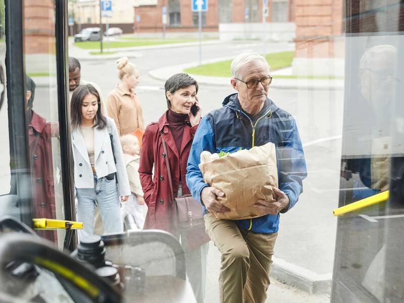 Der vorderer Einstieg eines Busses aus Sicht des Fahrersitzes. Ein älterer Herr mit einer Papiertüte in den Händen steigt gerade ein. Dahinter warten eine telefonierende Frau, drei weitere Passagiere und ein Kind. 