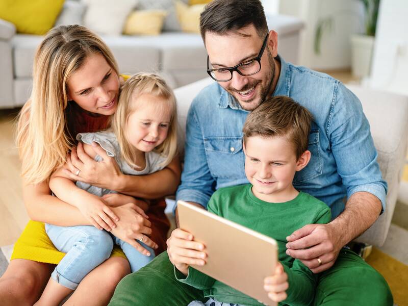 Eine Familie sitzt auf einem Teppich vor einem Sofa. Die Frau links umarmt ein kleines Mädchen. Rechts von ihr sitzt ein junge auf dem Schoss eines Mannes. Der Mann hält ein Tablett in der Hand, auf das alle schauen.