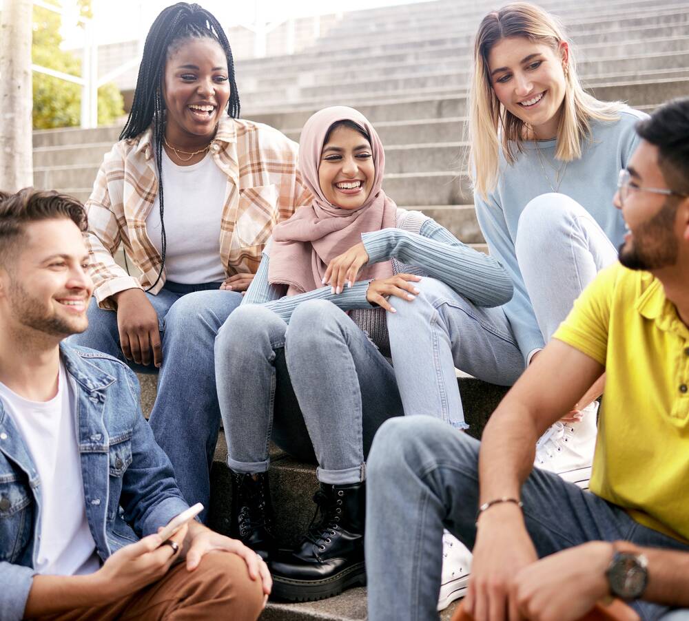 Drei junge Frauen sitzen im Freien auf einer Treppe zusammen. Auf den Stufen darunter sitzen zwei junge Männer. Sie lachen und schauen sich an.