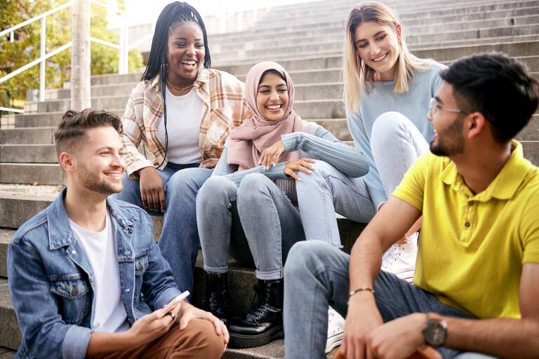 Drei junge Frauen sitzen im Freien auf einer Treppe zusammen. Auf den Stufen darunter sitzen zwei junge Männer. Sie lachen und schauen sich an.