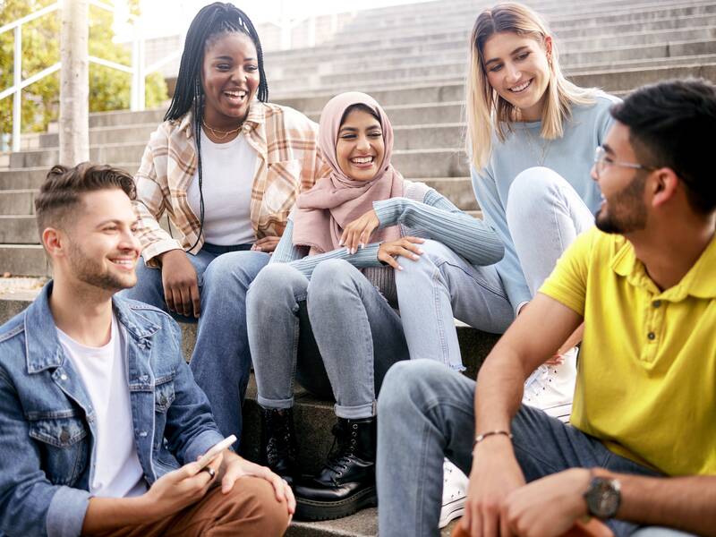 Drei junge Frauen sitzen im Freien auf einer Treppe zusammen. Auf den Stufen darunter sitzen zwei junge Männer. Sie lachen und schauen sich an.