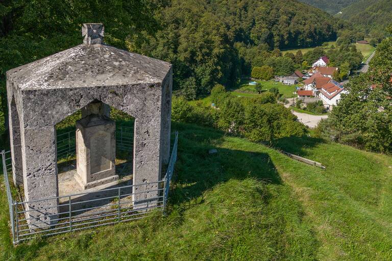 Im Vordergrund ein Pavillion aus Stein mit einer Steinstehle auf einem Hügel, dahinter Blick in die Landschaft mit einer kleinen Ortschaft und Wäldern.