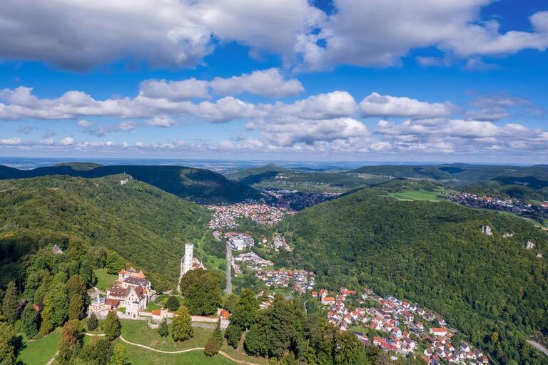 Blick von oben auf Schloss Lichtenstein, die Häuser im Tal sowie blauer Himmel mit weißen Wolken.