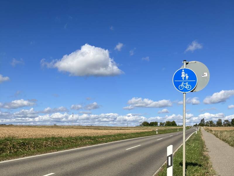 Eine Landstraße mit einem Fuß- und Radweg rechts daneben. Am Weg steht ein blaues Schild.