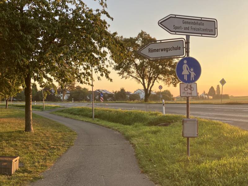 Ein Rad- und Fußweg in der Dämmerung. Links steht ein Baum, rechts ein Straßenschild.