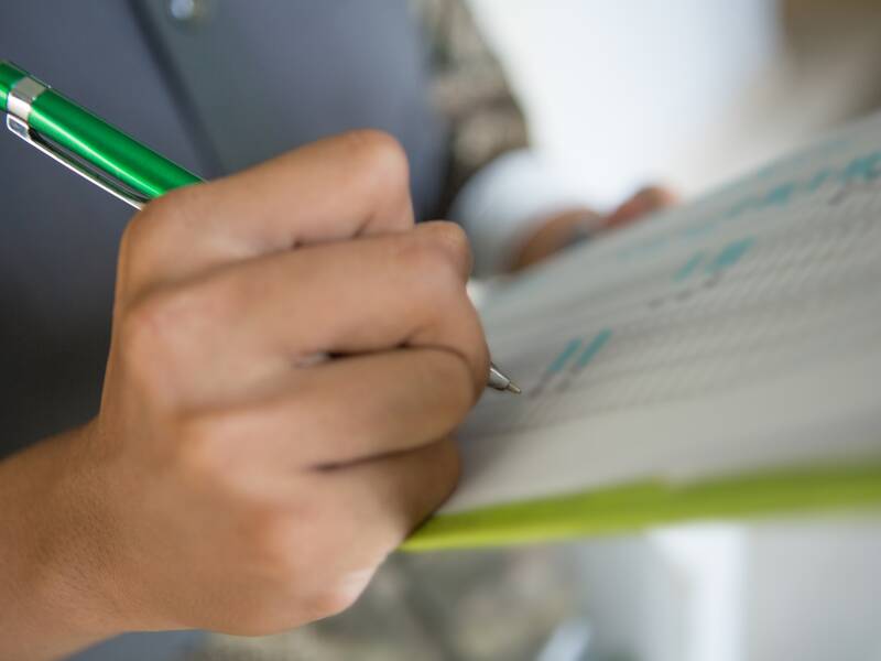 Ein grünes Klemmbrett mit Papier. Man sieht eine Hand, die einen grünen Stift hält und etwas auf dem Papier notiert.