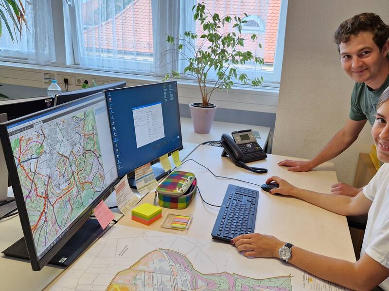 Zwei Personen lächeln in einem Büro, während sie an einem Schreibtisch mit Computern und Landkarten arbeiten.