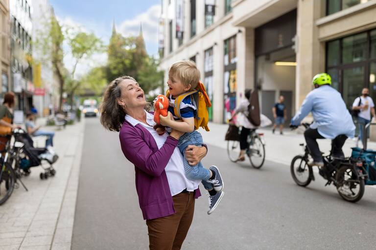 Eine ältere Frau mit lila Jacke hebt ein kleines Kind hoch. Sie steht in einer belebten Stadt. Daneben fahren Radfahrer vorbei.