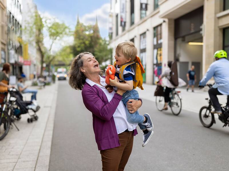 Eine ältere Frau mit lila Jacke hebt ein kleines Kind hoch. Sie steht in einer belebten Stadt. Daneben fahren Radfahrer vorbei.