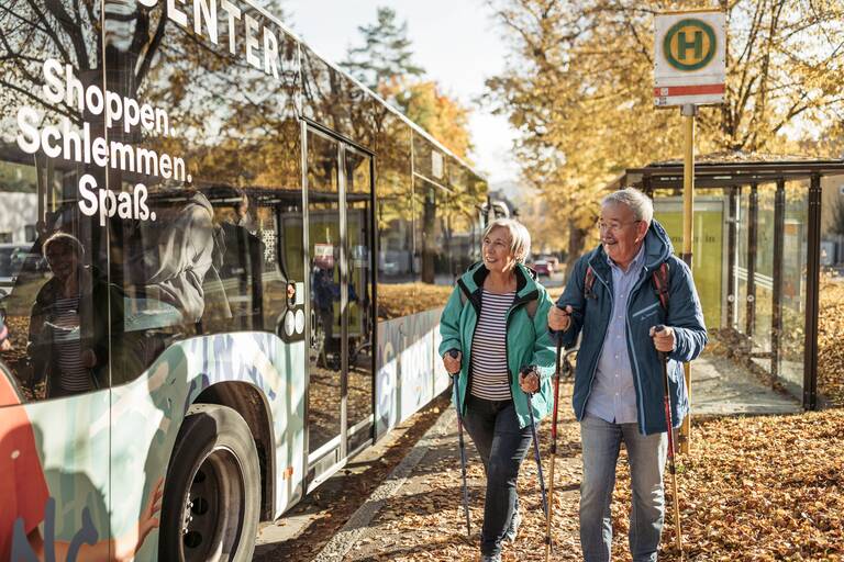 Eine Seniorin und ein Senior mit Wanderstöcken stehen an einer Bushaltestelle, an der ein Bus hält. Auf dem Boden liegen bunte Blätter.