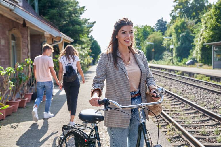 Eine junge Frau in einem Blazer steht mit einem Holland-Rad an einem Bahnsteig.