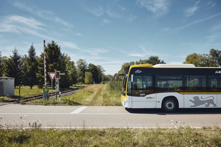 Ein schwarz-gelb-weißer Bus hält auf einer Straße vor einem Bahnübergang.