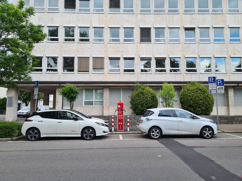 Zwei Elektro-Autos stehen an einer Ladesäule vor einem großen Gebäude.