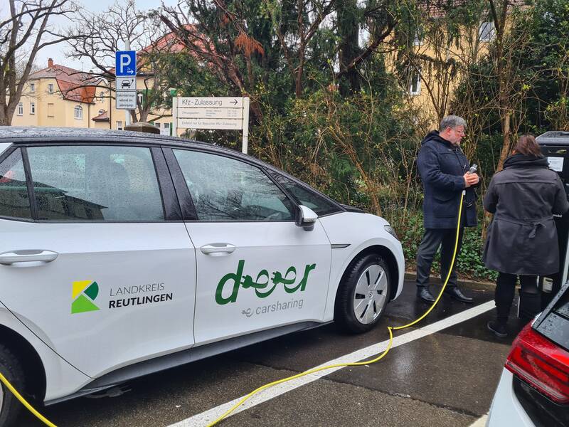 Ein E-Car Auto steht an einem Parkplatz. Rechts daneben sind ein Mann und eine Frau. Der Mann hält das Ladekabel in der Hand.