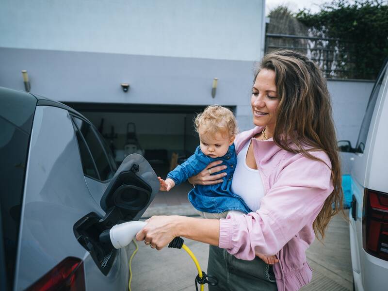 Eine Mutter mit Kind auf dem Arm lädt ihr E-Auto.