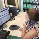 Eine junge Frau sitzt am Schreibtisch und telefoniert mit dem Handy. Auf dem Computer-Bildschirm vor ihr sind Fahrpläne aufgerufen.