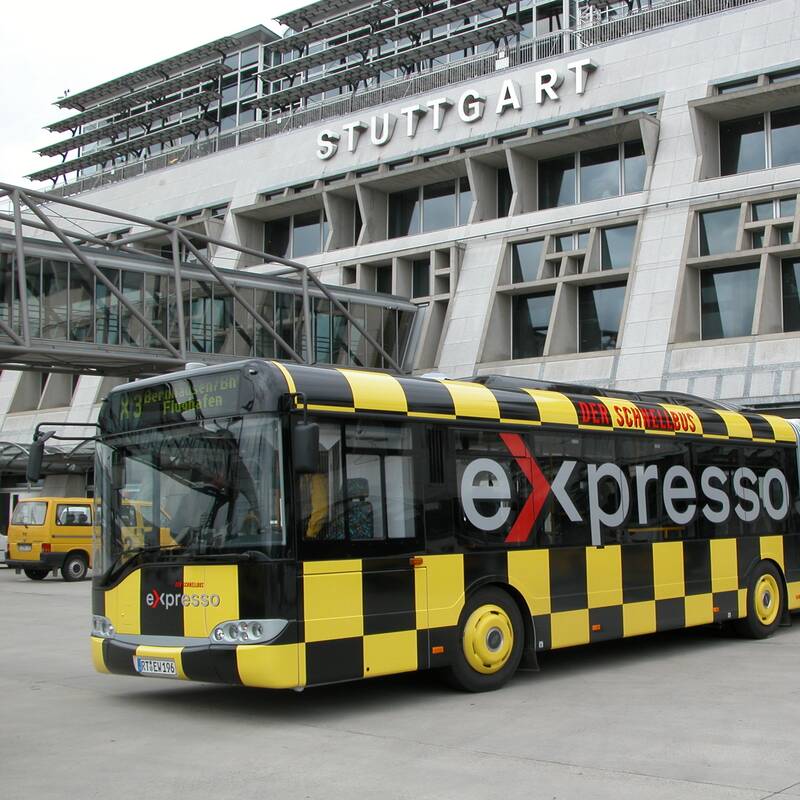 Ein schwarz-gelb karierter Bus mit roter Schrift steht vor einem Gebäude des Flughafens Stuttgart.