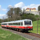 Ein historischer weiß-roter Zug fährt durch eine Landschaft. Oben im Bild auf einem Berg ist ein schlossähnliches Gebäude.