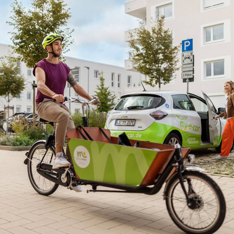 Ein Mann fährt in einem Wohngebiet ein Lastenfahrrad. Hinter ihm steht ein Elektroauto auf das eine Frau zugeht.