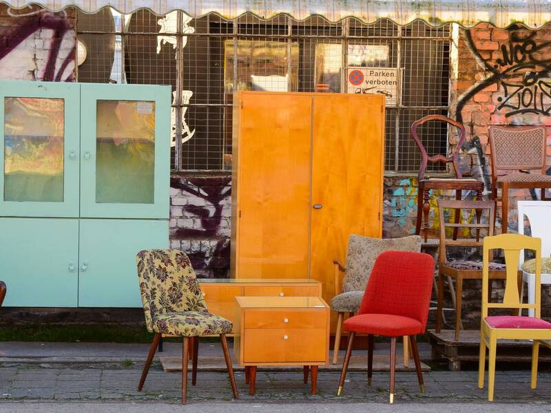 Viele verschieden Stühle aufgestellt. Ein grüner Schrank und ein brauner Schrank sind im Hintergrund. Man sieht im Hintergrund ein Gebäude mit Graffiti.