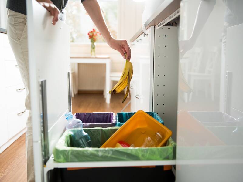 Man sieht eine Schublade. In der Schublade sind verschiedene Eimer mit einem grünem, einem violetten und einem blauen Sack. Eine Person schmeißt eine Bananenschale in den Eimer.