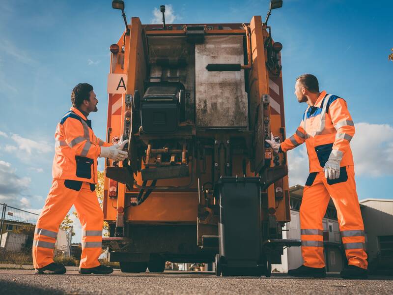 Zwei Personen in orangener Arbeitskleidung stehen neben einem orangenen Müllwagen. Eine schwarze Tonne wird in den Müllwagen geleert.