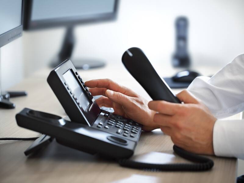 Ein schwarzes Telefon steht auf einem Tisch. Eine Person hat den Hörer des Telefons in der Hand.