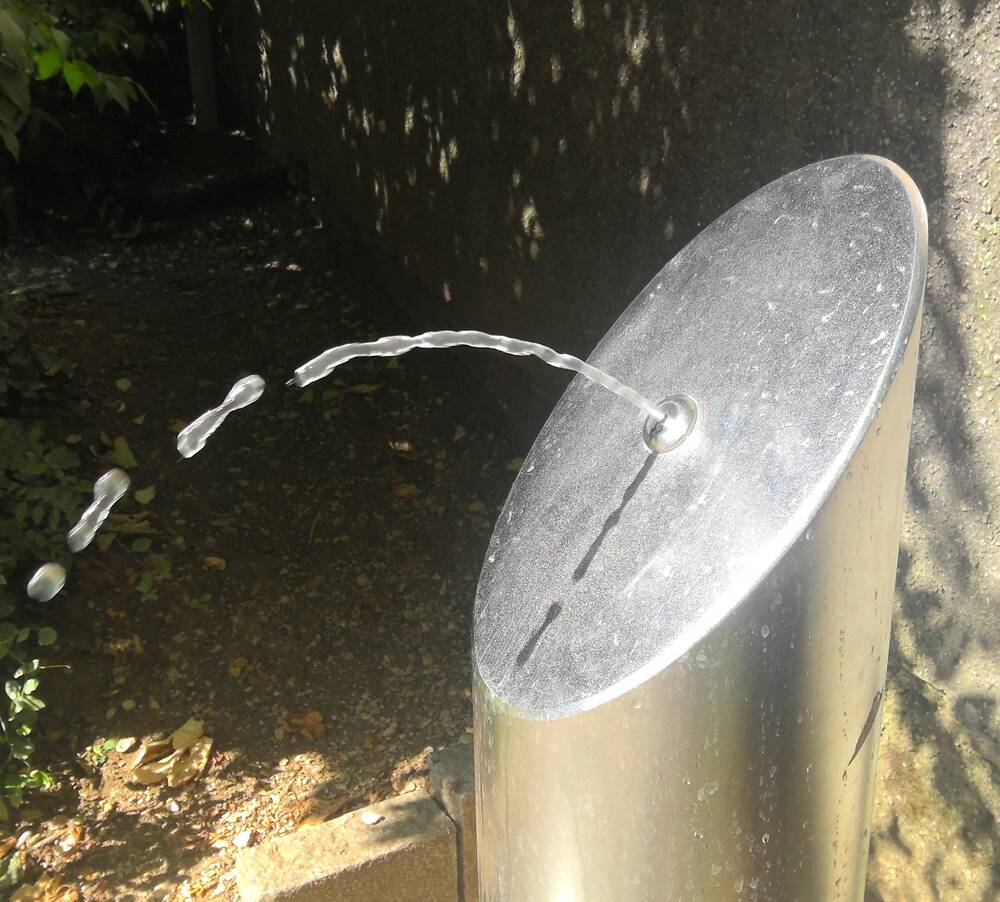 Aus einem öffentlicher Trinkwasserspender aus Metall sprudelt Wasser. Im Hintergrund eine Mauer und Pflanzen.