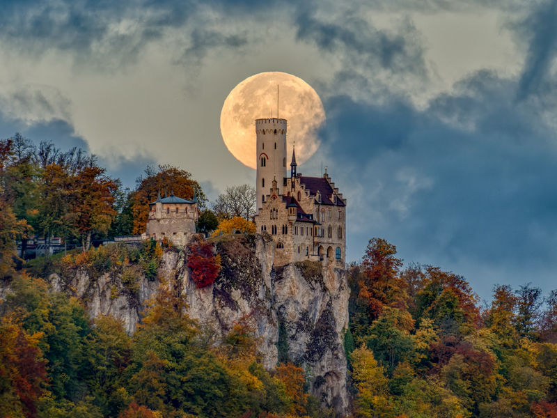 Eine Burg auf einem Hügel und einem wolkenbehangenen Himmel. Hinter dem Burgturm ist der Vollmond.