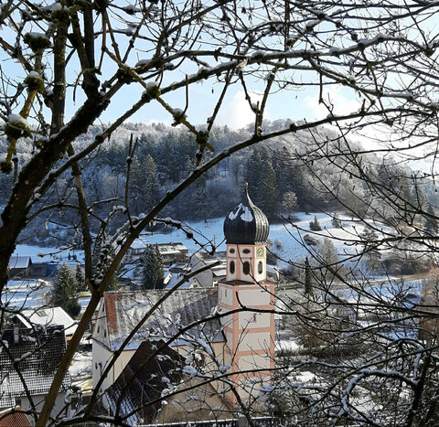 Blick auf einen verschneiten Kirchturm und ein Dorf in hügeliger Landschaft, eingerahmt von schneebedeckten Ästen.