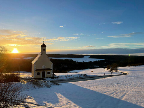 Eine kleine Kapelle steht in einer verschneiten Landschaft bei Sonnenuntergang, mit einem gefrorenen See und bewaldete Hügel im Hintergrund.