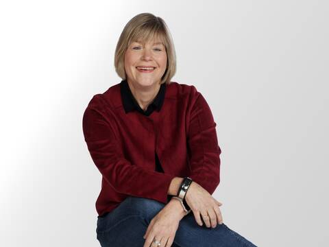 Ein Portrait einer Frau mit kurzen blonden Haaren. Sie lächelt und trägt ein schwarzes Hemd, eine rote Jacke und eine blaue Jeans.