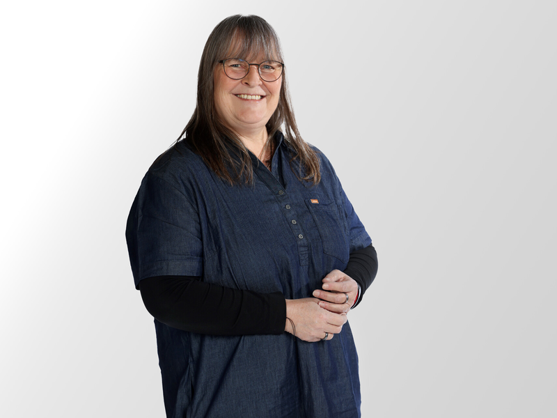 Ein Portrait einer Frau mit schulterlangen braunen Haaren. Sie trägt eine Brille und lächelt. Sie trägt ein langärmliges schwarzes Shirt und eine dunkelblaue Bluse.