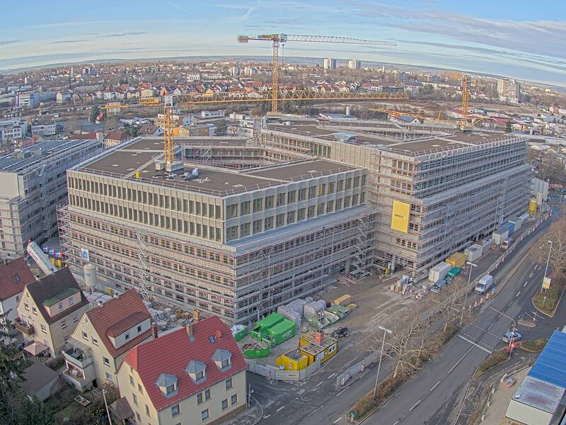 Das Bild zeigt eine Baustelle eines großen Gebäudekomplexes in einer städtischen Umgebung mit Kränen und umliegenden Wohnhäusern.